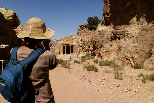 A person wearing a wide-brimmed hat and blue backpack photographs ancient rock-cut architecture amidst a rugged desert landscape, with sandy pathways and towering cliffs surrounding them.